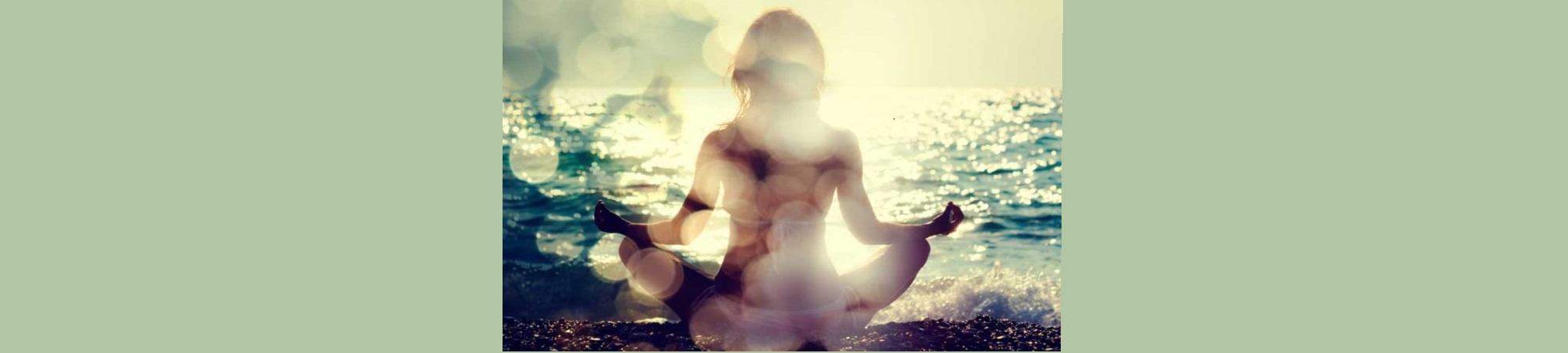 Meditating woman in yoga pose, facing away toward sunlight on ocean water. 